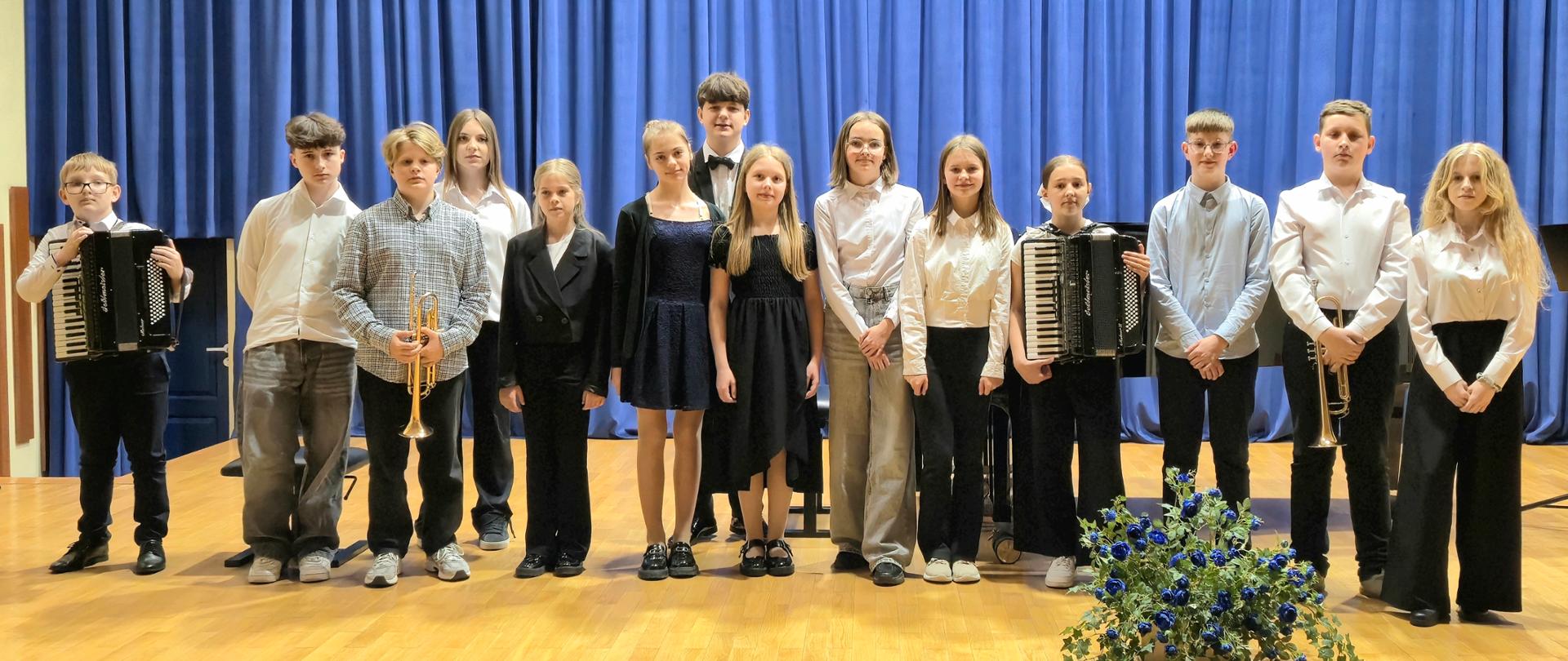 grupa dzieci w galowych strojach na scenie, niektórzy trzymają instrumenty, za nimi niebieska kotara