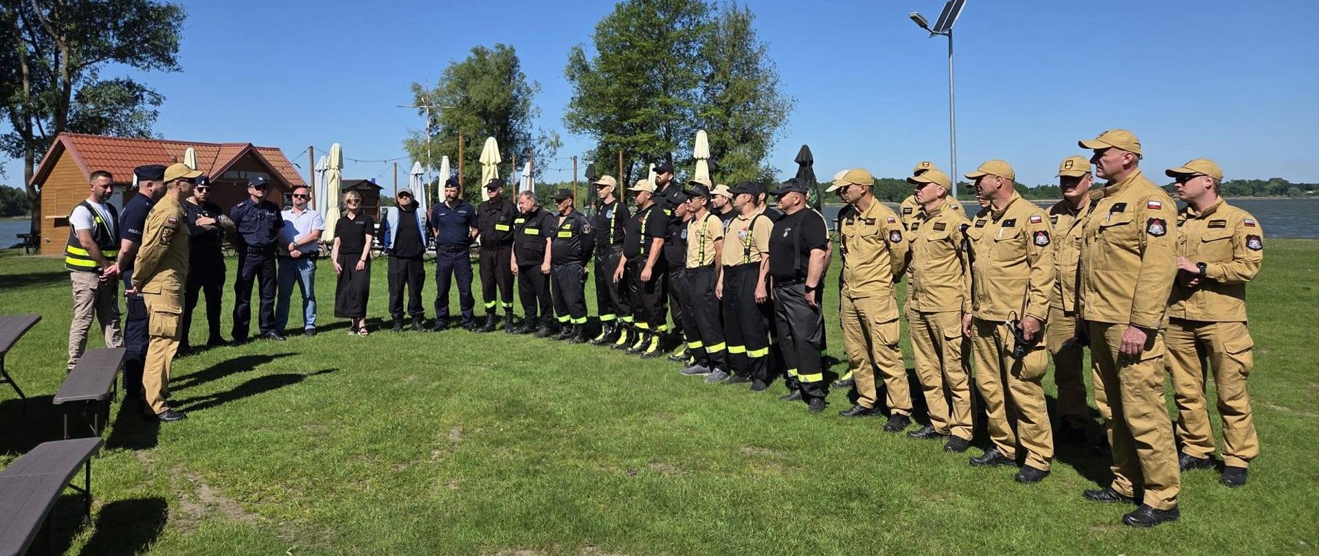 Uczestnicy ćwiczeń – strażacy PSP, druhowie OSP, policjanci oraz przedstawiciele instytucji współpracujących – stoją podczas odprawy organizacyjnej na terenie ośrodka nad Jeziorem Gopło.