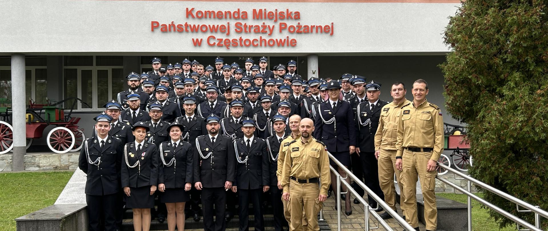 Na zdjęciu widać dużą grupę strażaków pozujących do wspólnego zdjęcia przed budynkiem Komendy Miejskiej Państwowej Straży Pożarnej w Częstochowie. Większość osób ubrana jest w galowe, ciemne mundury strażackie z czapkami, a kilku strażaków z przodu ma mundury w kolorze piaskowym. Grupa stoi na schodach i podjeździe prowadzącym do wejścia budynku. Nad nimi widoczny jest napis z nazwą komendy. Po lewej stronie częściowo widać starą sikawkę konną, a po prawej rośnie drzewo iglaste. Scena ma charakter pamiątkowy i formalny.