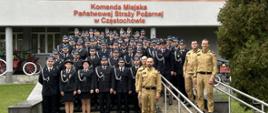 Na zdjęciu widać dużą grupę strażaków pozujących do wspólnego zdjęcia przed budynkiem Komendy Miejskiej Państwowej Straży Pożarnej w Częstochowie. Większość osób ubrana jest w galowe, ciemne mundury strażackie z czapkami, a kilku strażaków z przodu ma mundury w kolorze piaskowym. Grupa stoi na schodach i podjeździe prowadzącym do wejścia budynku. Nad nimi widoczny jest napis z nazwą komendy. Po lewej stronie częściowo widać starą sikawkę konną, a po prawej rośnie drzewo iglaste. Scena ma charakter pamiątkowy i formalny.
