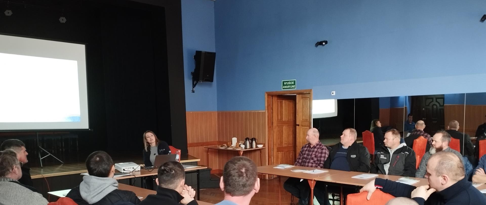  Uczestnicy siedzą przy stołach, w głębi siedzi prowadząca szkolenie, za jej plecami jest ekran