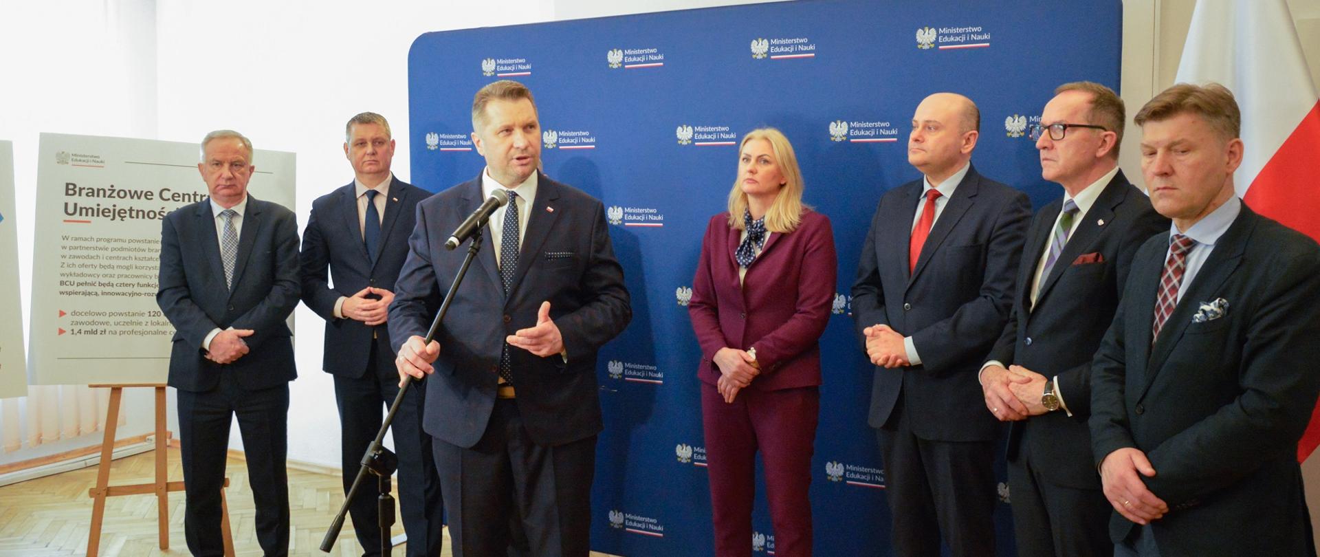 Przed niebieską ścianką stoi grupa mężczyzn w garniturach i jasnowłosa kobieta w ciemnoczerwonym ubraniu, przed nimi minister Czarnek mówi do mikrofonu na stojaku, z tyłu stoi tablica z napisem Branżowe Centra Umiejętności, za nią biała ściana i zasłonięte białymi roletami okno.