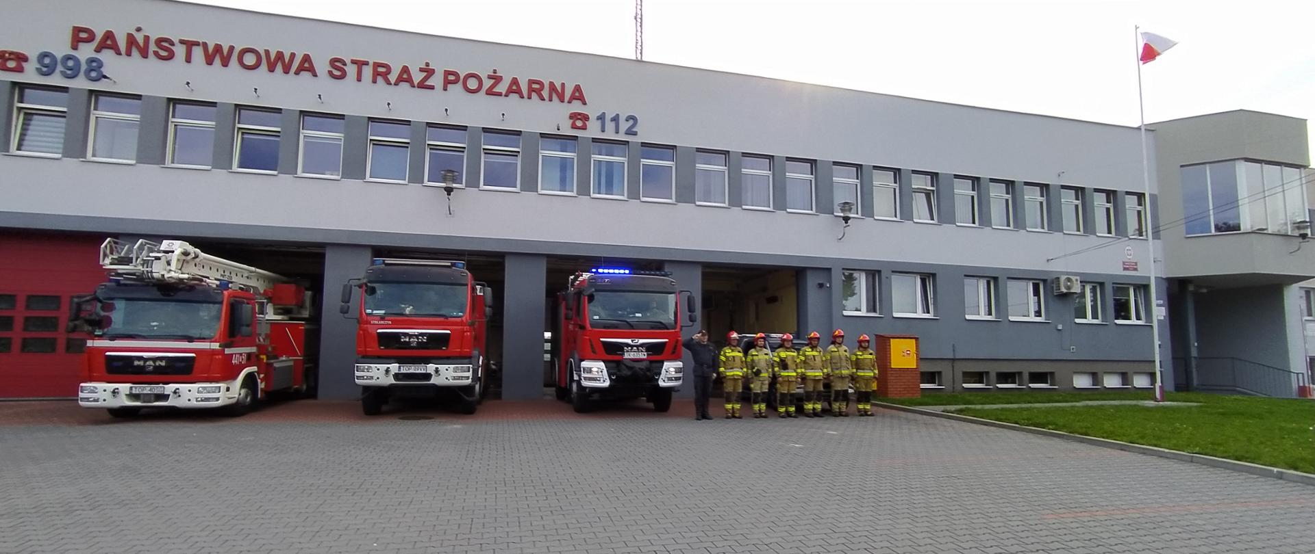 Zdjęcie przedstawia strażaków oddających honory przed remizą KP PSP. W tle widać budynek oraz samochody strażackie