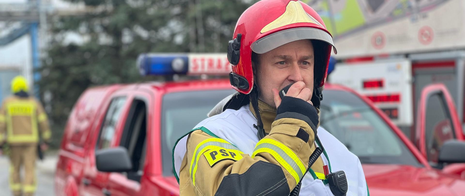 Strażak w czerwonym hełmie i biało-zielonej kamizelce na tle czerwonego samochodu.