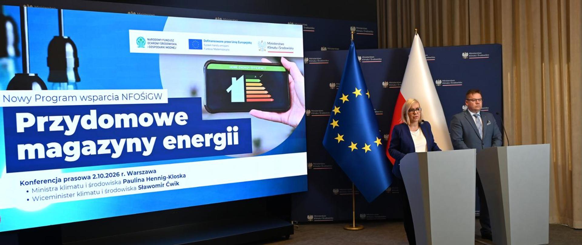 Conference room with a large presentation screen displaying the title “Residential Energy Storage Systems” and an image of a building energy efficiency rating shown on a smartphone. Speakers stand at podiums beside the screen. Polish and European Union flags and a backdrop of the Ministry of Climate and Environment are visible in the background.