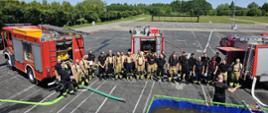 Zdjęcie przedstawia grupę strażaków OSP i PSP stojących tyłem do samochodów pożarniczych. Na pierwszym planie umiejscowiony jest przenośny zbiornik na wodę oraz prowadzący zajęcia.