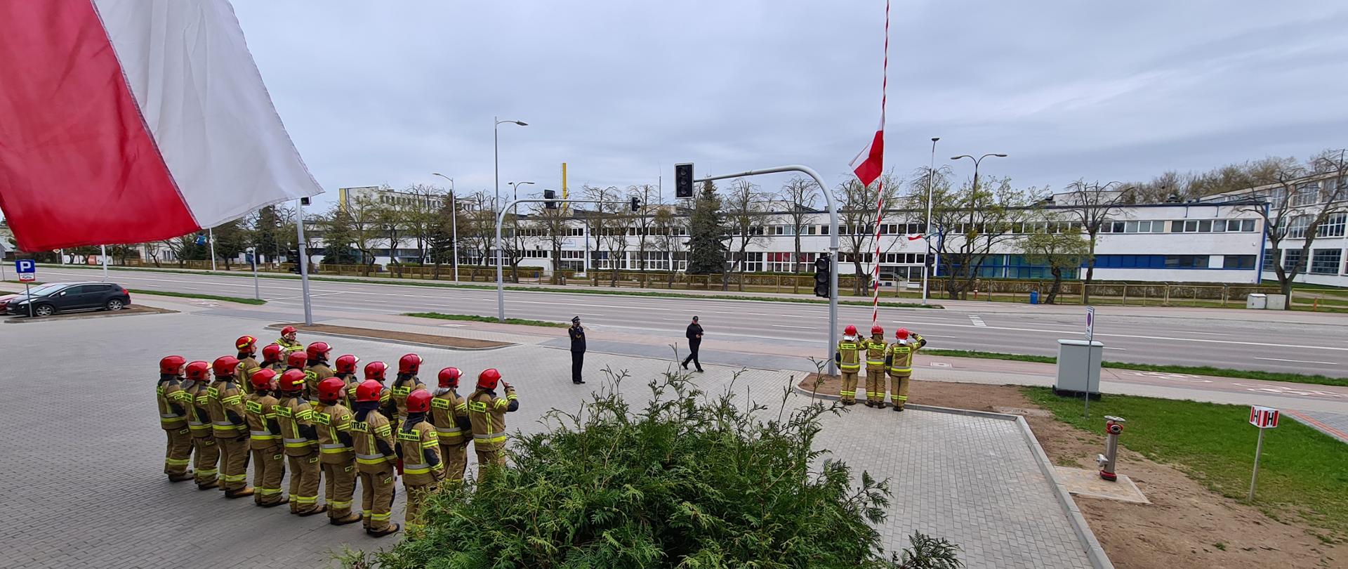 Zdjęcie przedstawia pododdziały KP PSP w Ełku podczas uroczystości Obchodów Dnia Flagi Rzeczypospolitej Polskiej, na zdjęciu widać strażaków ustawionych w dwu szeregu frontem do masztu flagowego, poczt flagowy przy maszcie flagowym oraz dowódcę uroczystości