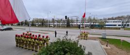 Zdjęcie przedstawia pododdziały KP PSP w Ełku podczas uroczystości Obchodów Dnia Flagi Rzeczypospolitej Polskiej, na zdjęciu widać strażaków ustawionych w dwu szeregu frontem do masztu flagowego, poczt flagowy przy maszcie flagowym oraz dowódcę uroczystości - kadr panoramiczny