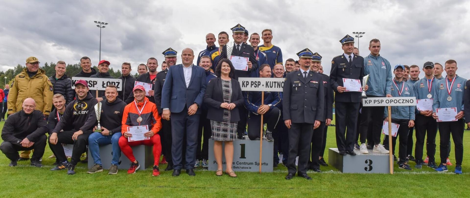 trzy drużyny strażaków stojące na i obok stopni tworzących podium, każda drużyna trzyma tabliczkę ze skrótem komendy którą reprezentują, podium ustawione jest na zielonej murawie