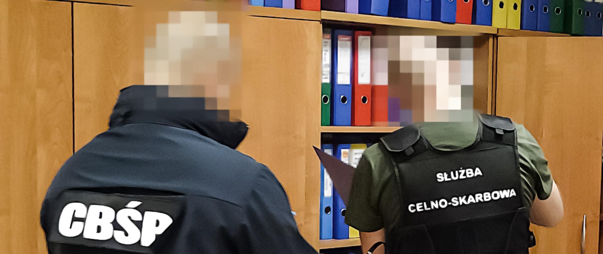 Dwóch funkcjonariuszy służb w odzieży z oznaczeniami „CBŚP” oraz „Służba Celno-Skarbowa” stoi w biurze przed regałem z kolorowymi segregatorami i przegląda dokumenty. 