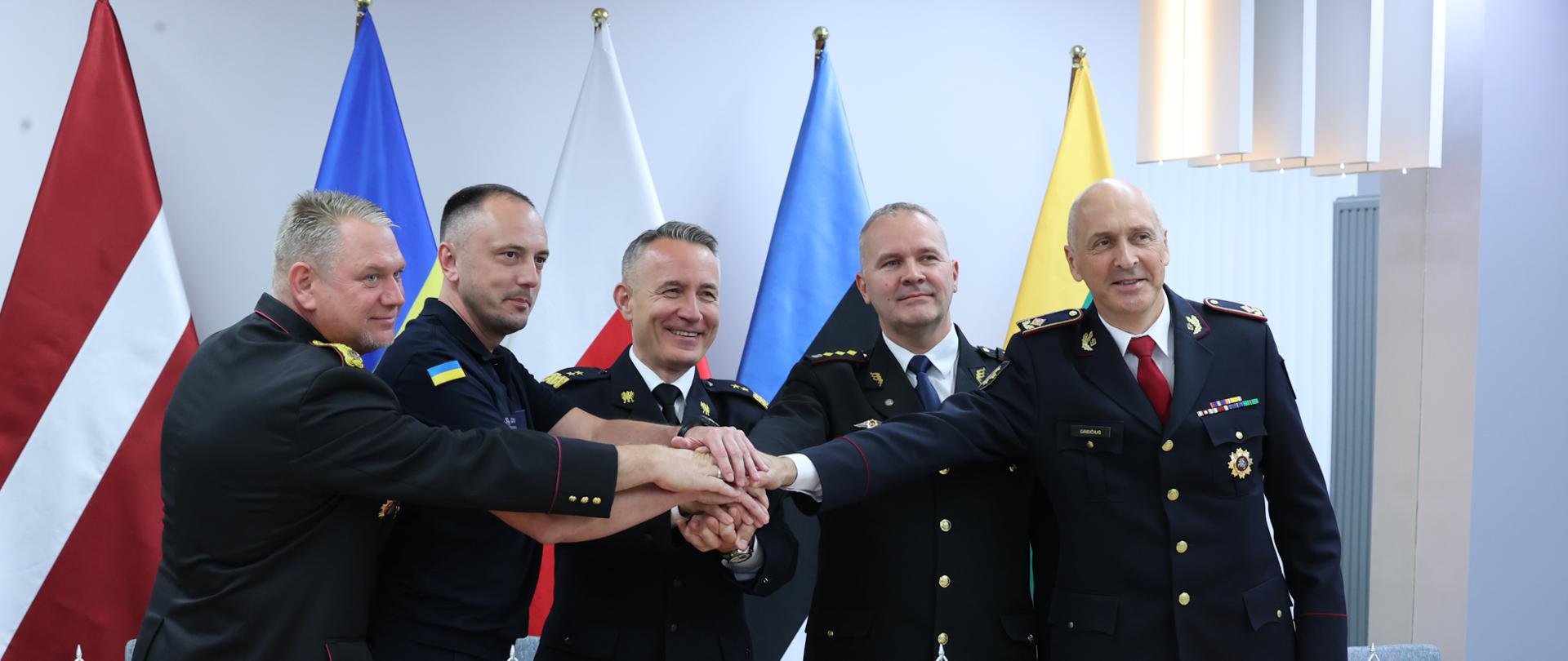 Na zdjęciu widać Szefów Służb Ratowniczych Ukrainy, Litwy, Łotwy, Estonii i komendanta głównego PSP którzy nakładają na siebie swoje dłonie wyrażając jedność. W tle flagi.