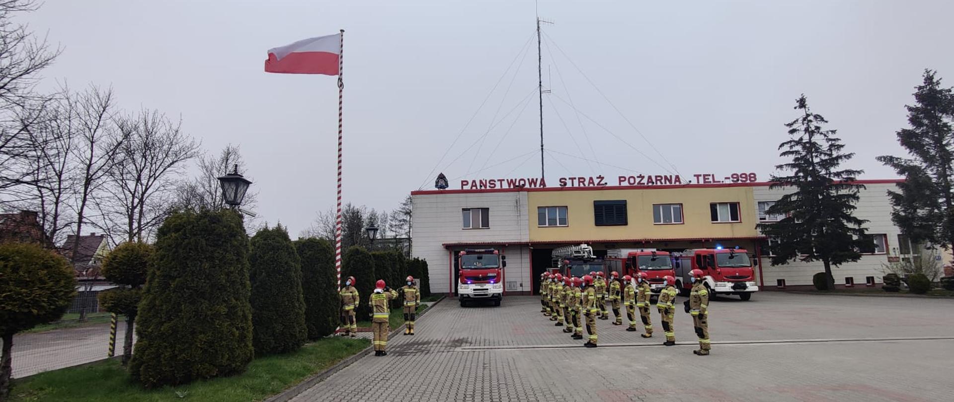 Zbiórka strażaków przez budynkiem Komendy podczas uroczystego wciągnięcia flagi na maszt. W tle pojazdy pożarnicze w z włączonymi światłami błyskowymi. 