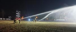 Nocne manewry z taktyki zwalczania pożarów z dostarczania wody na duże odległości dla słuchaczy szkolenia podstawowego w zawodzie strażak, dwie grupy strażaków ustawione w pewnej odległości od siebie na murawie boiska piłkarskiego. Jeden ze strażaków w każdej grupie trzyma prądownicę z której na dużą odległość leci strumień wody