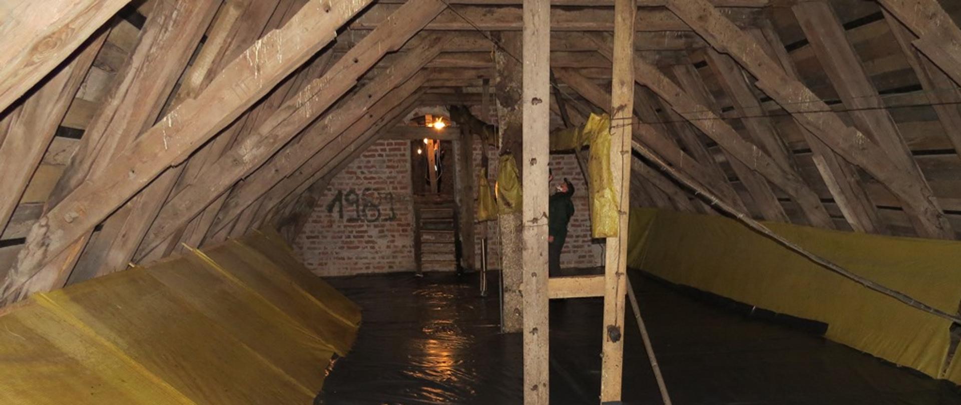 Oświetlony strych kościoła i podłoga zabezpieczona czarną folią, w tle widać człowieka wykonującego prace. 