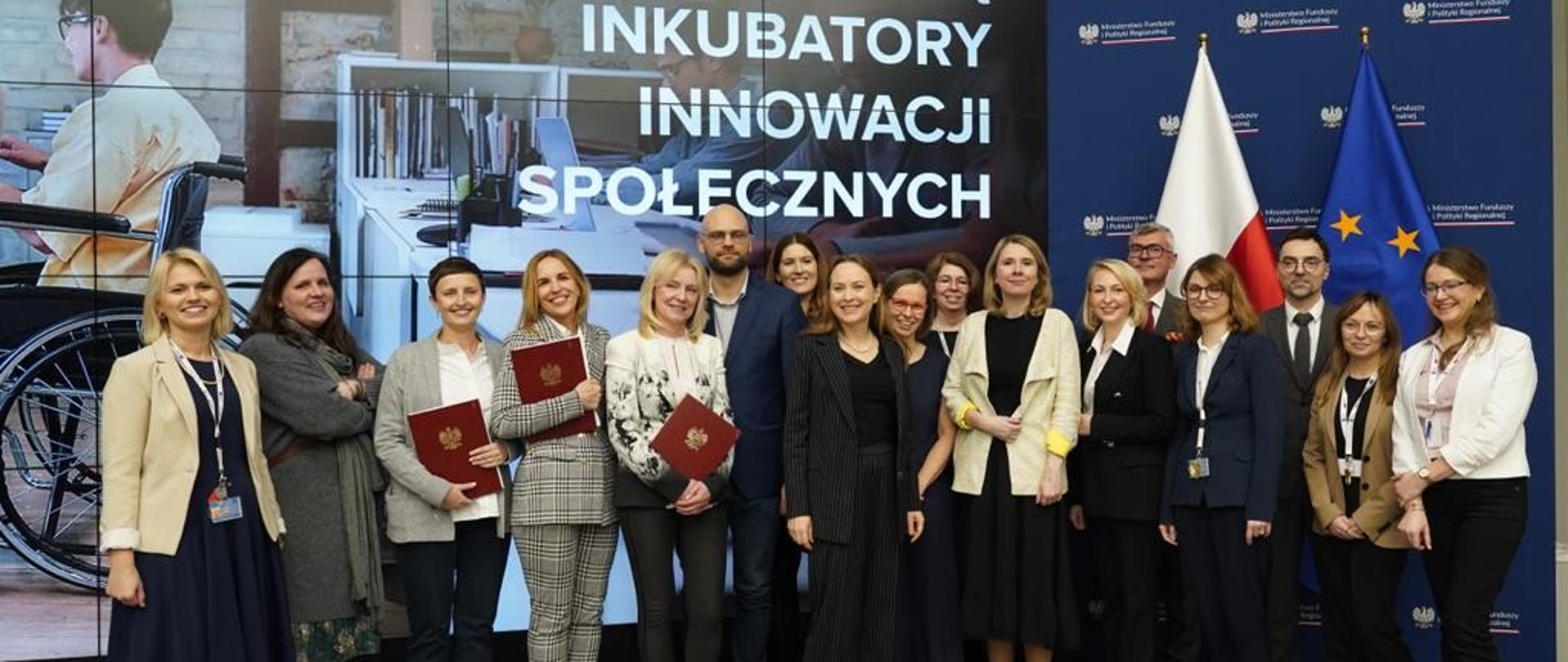 Minister funduszy i polityki regionalnej Katarzyna Pełczyńska-Nałęcz i wiceministra Monika Sikora z beneficjenci konkursu na inkubatory na innowacje społeczne, uczestnicy stoją na tle zdjęcia przedstawiającego młodego mężczyznę na wózku inwalidzkim