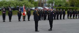 Na zdjęciu widoczny Dowódca uroczystości starszy kapitan Kacper Węgrzyn w mundurze galowym składający meldunek Zastępy Opolskiego Komendanta Wojewódzkiego Państwowej Straży Pożarnej starszemu brygadierowi Arkadiuszowi Kuśnierskiemu w mundurze galowym. W tle widoczny pododdział pożarniczy Komendy Powiatowej Państwowej Straży Pożarnej w Głubczycach w dwuszeregu .