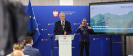 Wicepremier, minister rozwoju, pracy i technologii Jarosław Gowin stojący przy mównicy z mikrofonem, po jego prawej stronie stoi tłumacz języka migowego oraz ekran telewizora z wyświetloną grafiką przedstawiającą dwóch turystów w górach z plecakami i napisem TurystykApp - najlepsza droga do odkrywania usług turystycznych w Polsce.
