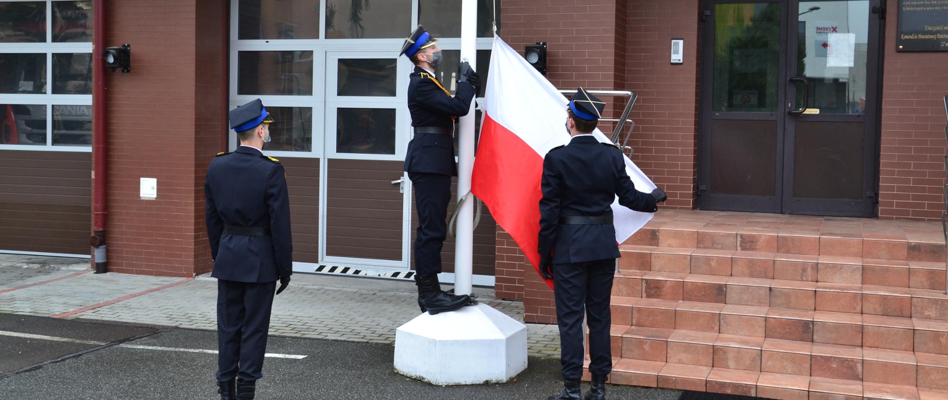 Poczet flagowy zapina flagę do masztu. D-ca pocztu w prawej ręce trzyma róg flagi, flagowy gotowy do podniesienia flagi państwowej. Strażacy ubrani w mundury wyjściowe.