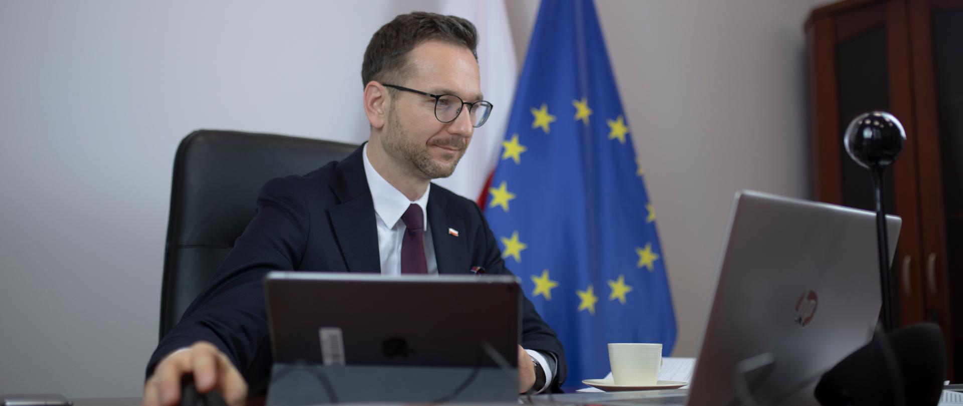 wiceminister Waldemar Buda przed komputerem, za nim flagi Polski i Unii Europejskiej