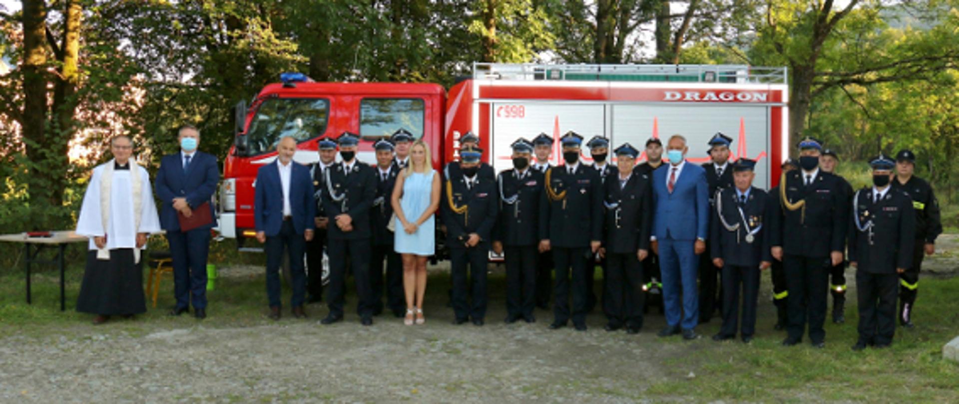 Na zdjęciu przedstawiciele władz, komendant wojewódzki oraz druhowie osp łodyna, na tle drzew samochód pożarniczy