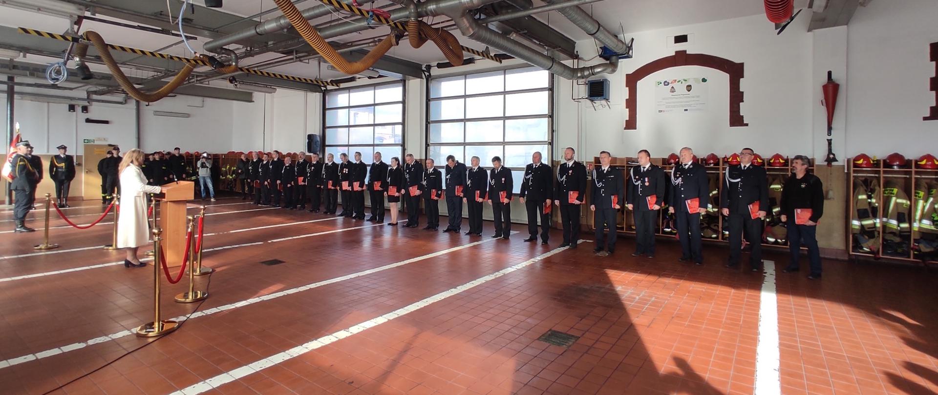 Zdjęcie przedstawia strażaków w mundurach stojących w garażu podczas uroczystej zbiórki. Po lewej stronie widoczna osoba przemawiająca przy mównicy