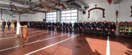 Zdjęcie przedstawia strażaków w mundurach stojących w garażu podczas uroczystej zbiórki. Po lewej stronie widoczna osoba przemawiająca przy mównicy