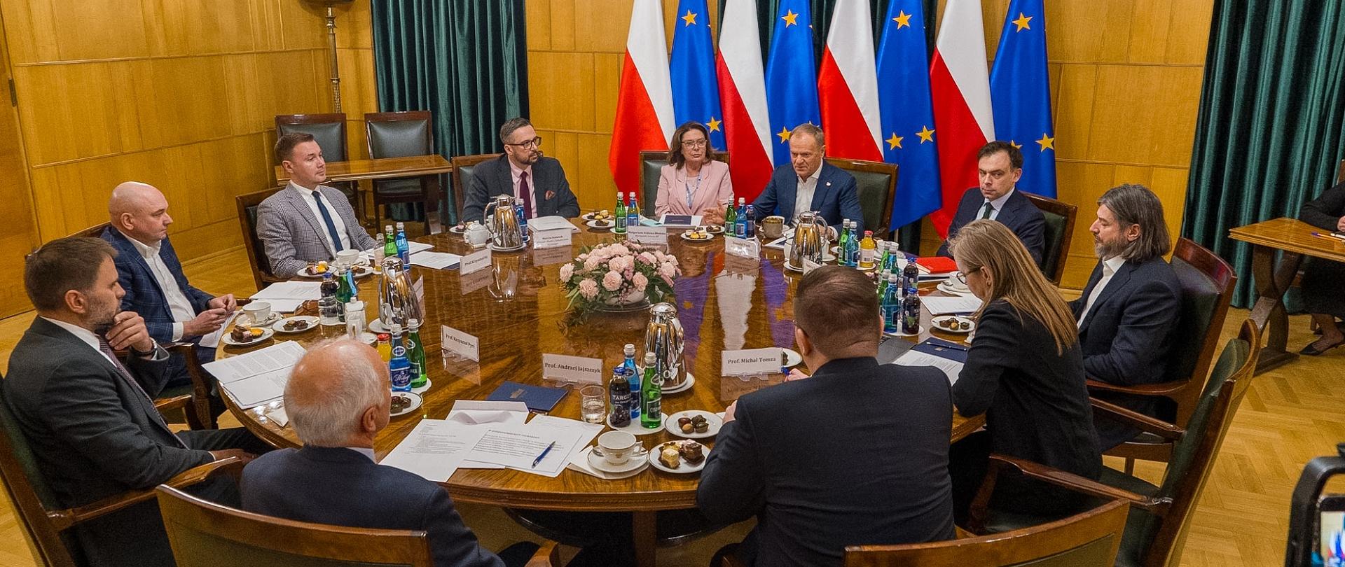 Zdjęcie z pewnej odległości, za okrągłym stołem siedzi kilkanaście osób, rozmawiają, na środku stołu różowe kwiaty, w tle pod żółtą ścianą rząd flag Polski i UE.