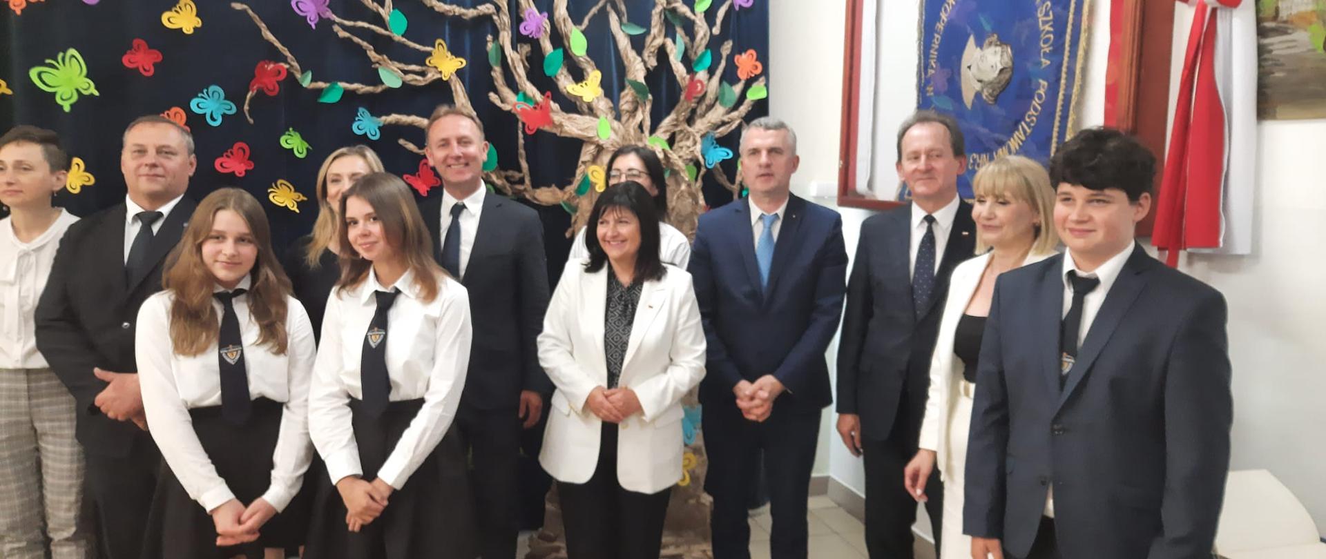 Wiceminister Włodzimierz Bernacki w gronie uczniów i nauczycieli szkoły. W tle dekoracja drzewo z kolorowymi motylami, sztandar szkoły i flaga biało-czerwona