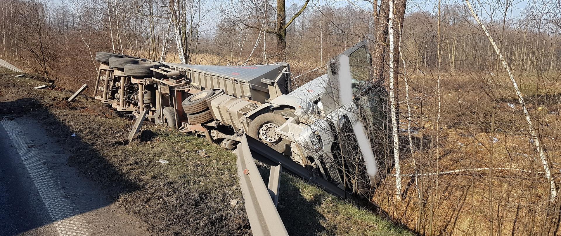 Samochód ciężarowy z naczepą leży na boku w rowie, w tle drzewa. Samochód koloru szarego. 