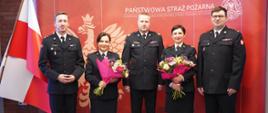 strażacy - kobiety z kwiatami i meżczyźni stoją obok siebie, w tle czerwona ścianka z napisem państwowa straż pożarna oraz biało czerwona flaga