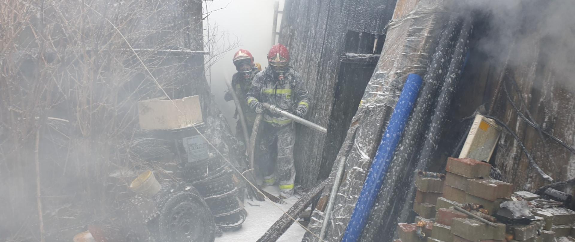 Strażacy ubrani w ubrania bojowe wchodzą do palącego się budynku gospodarczego. Strażacy i budynek są pokryci pianą ciężką gaśniczą. Budynek jest drewniany i obok niego w sąsiedztwie stoi kolejny budynek. Przy budynku leży złożona kostka brukowa oraz felgi samochodowe.