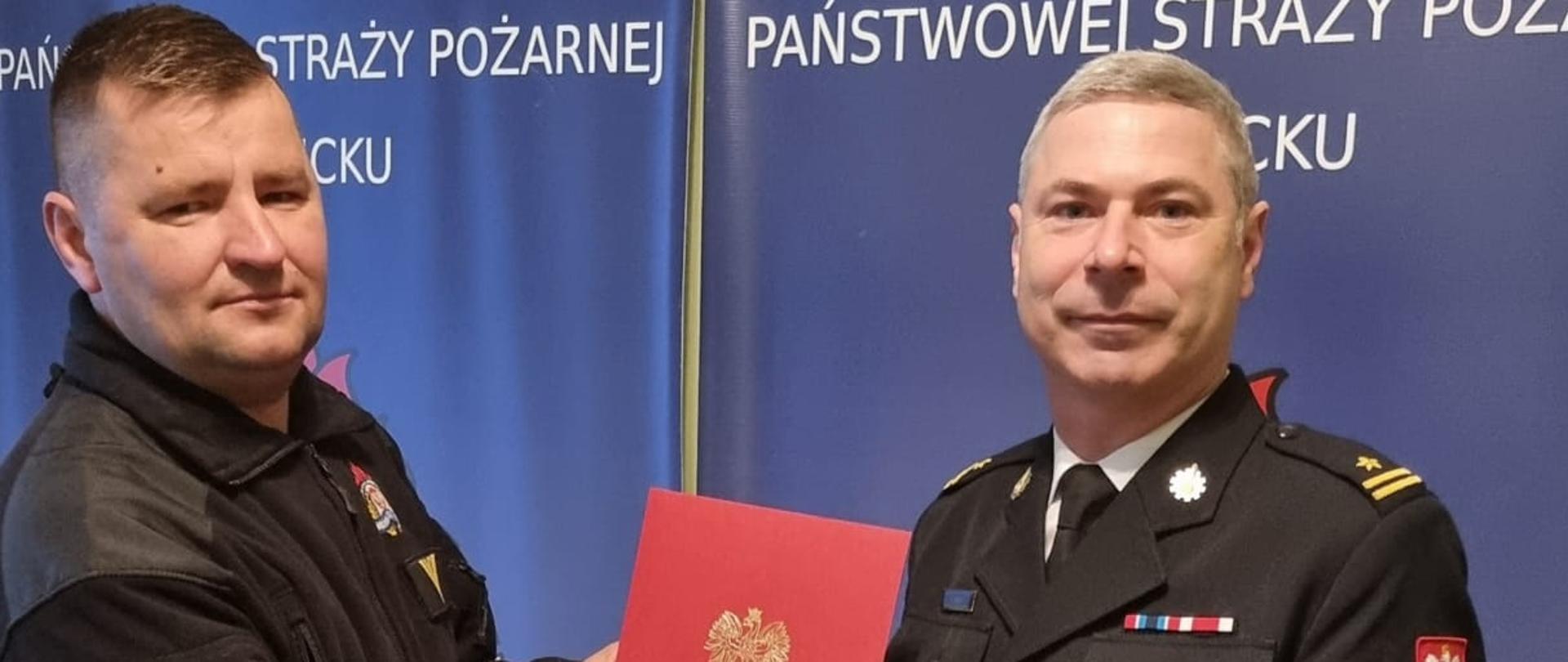 Na zdjęciu widać strażaków zastępca komendanta powiatowego w mundurze wyjściowym wręcza czerwoną teczkę z nagrodą strażakowi w mundurze koszarowym.