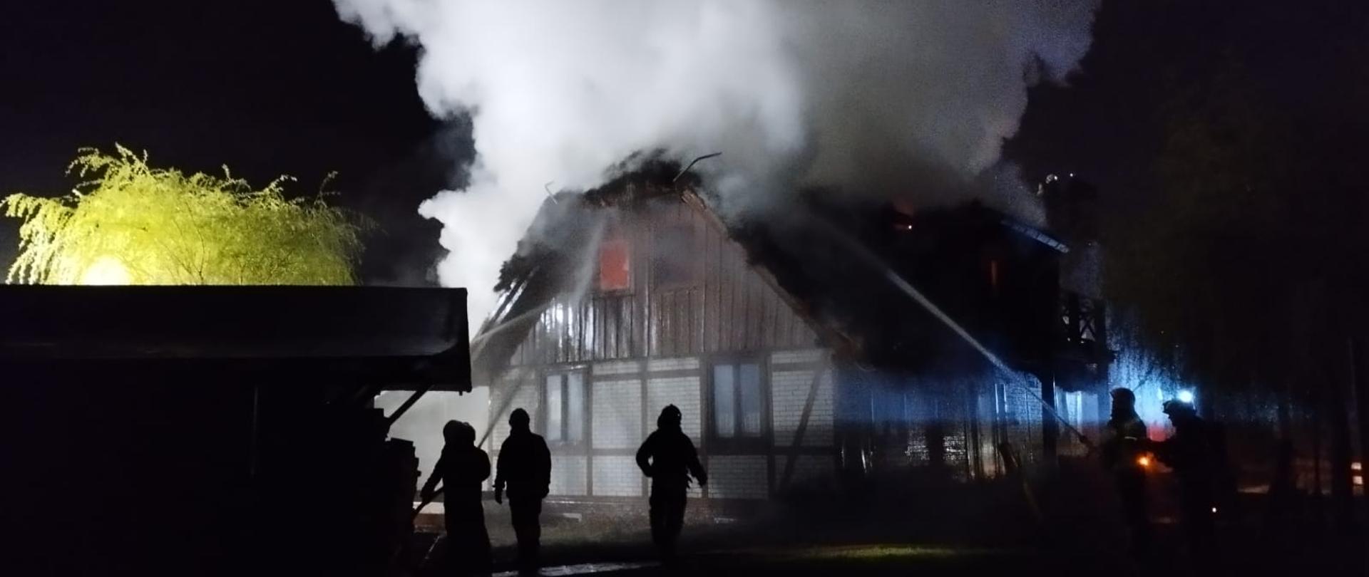 Akcja gaszenia domu. Jest noc, strażacy ze wszystkich stron podają prądy wody. Z dachu budynku wydobywają się kłęby dymu, w jednym z okien widoczny jest ogień.