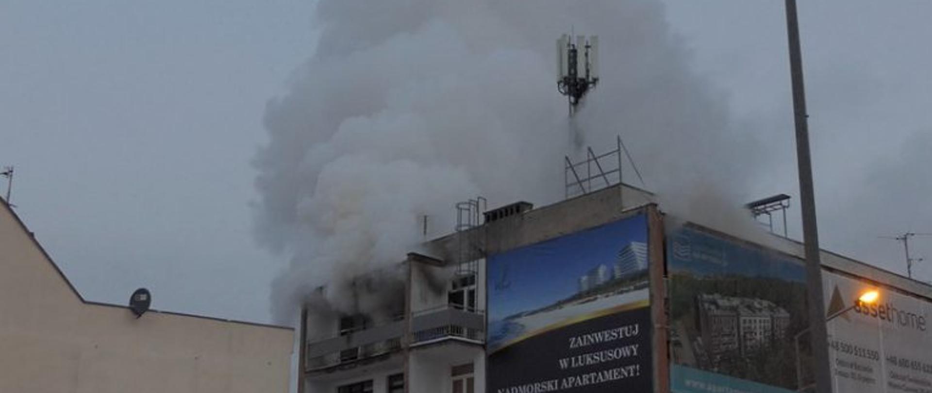 Zdjęcie przedstawia budynek 3 kondygnacyjny, na ostatniej kondygnacji wydobywa się dym.