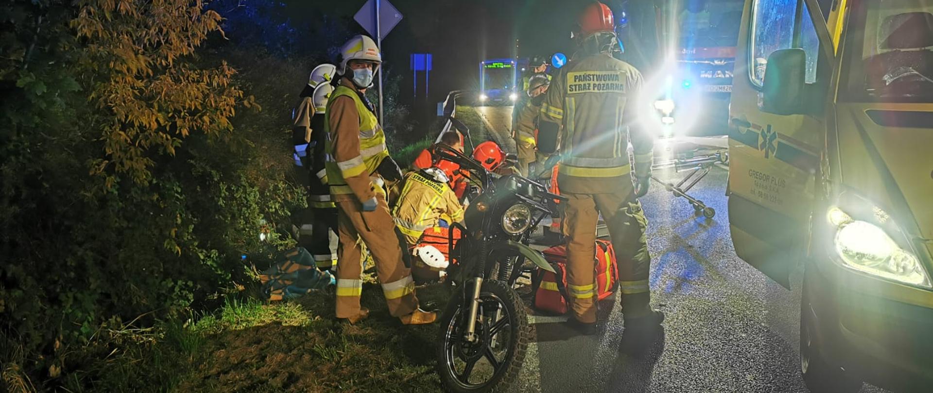 Na zdjęciu strażacy i ratownicy medyczni udzielają pomocy poszkodowanemu motocykliście, na poboczu motocykl na jezdni karetka pogotowia