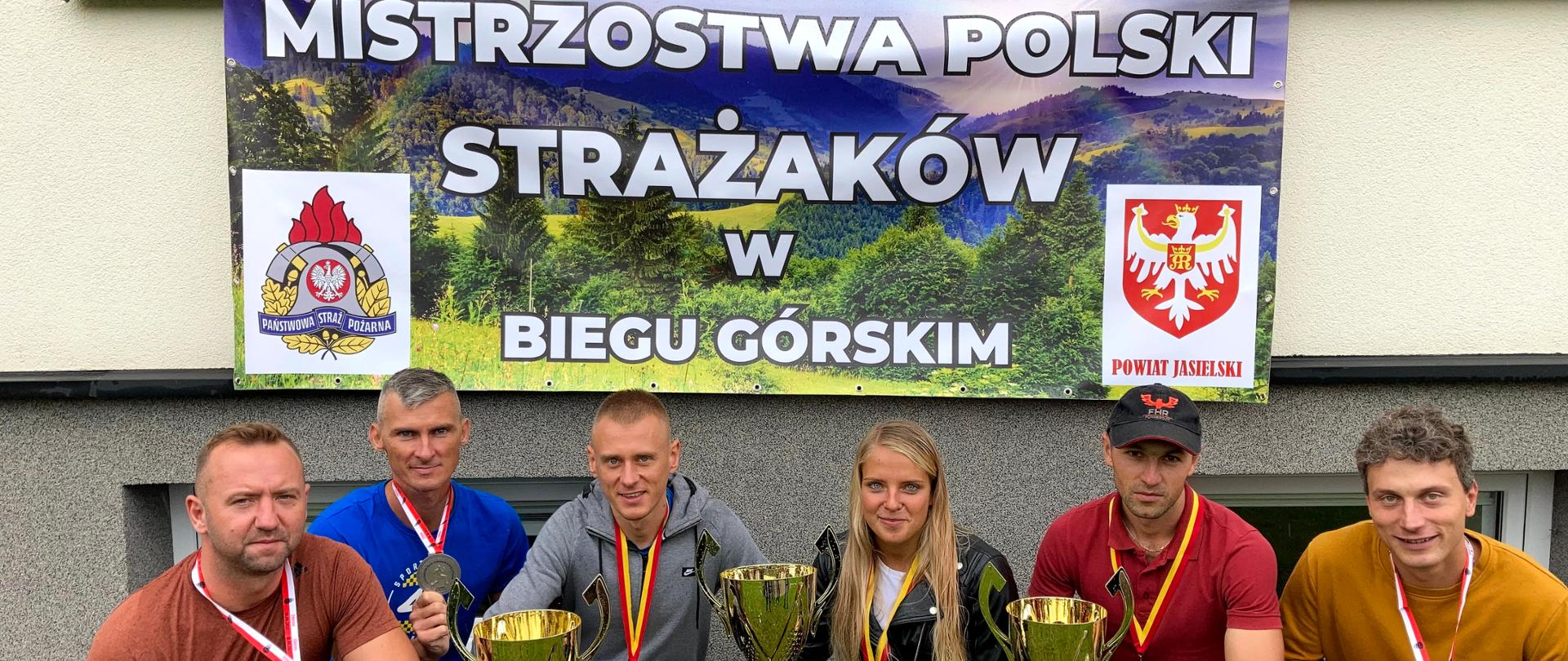 Na zdjęciu pozuje sześć osób (pięciu mężczyzn oraz jedna kobieta) z medalami i pucharami. Nad pozującymi wisi banner z logiem PSP, herbem powiatu oraz napisem Mistrzostwa Polski Strażaków w Biegu Górskim.