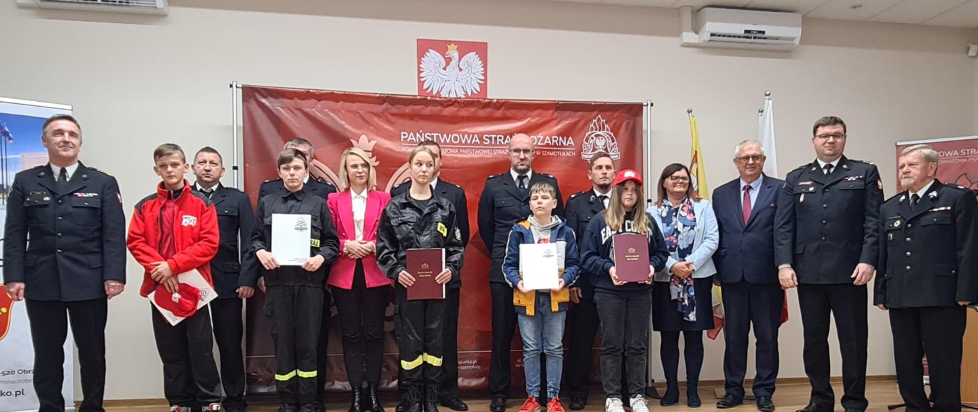 zdjęcie grupowe strażaków, osób cywilnych oraz dzieci, na czerwonym tle