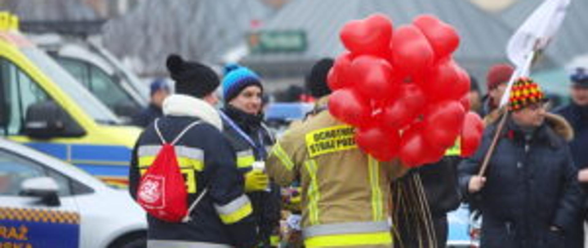 Na zimowym placu widać strażaków i uczestników akcji charytatywnej, z których jeden ubrany w strój OSP trzyma pęk czerwonych balonów, a w tle stoją ludzie oraz zaparkowany radiowóz.