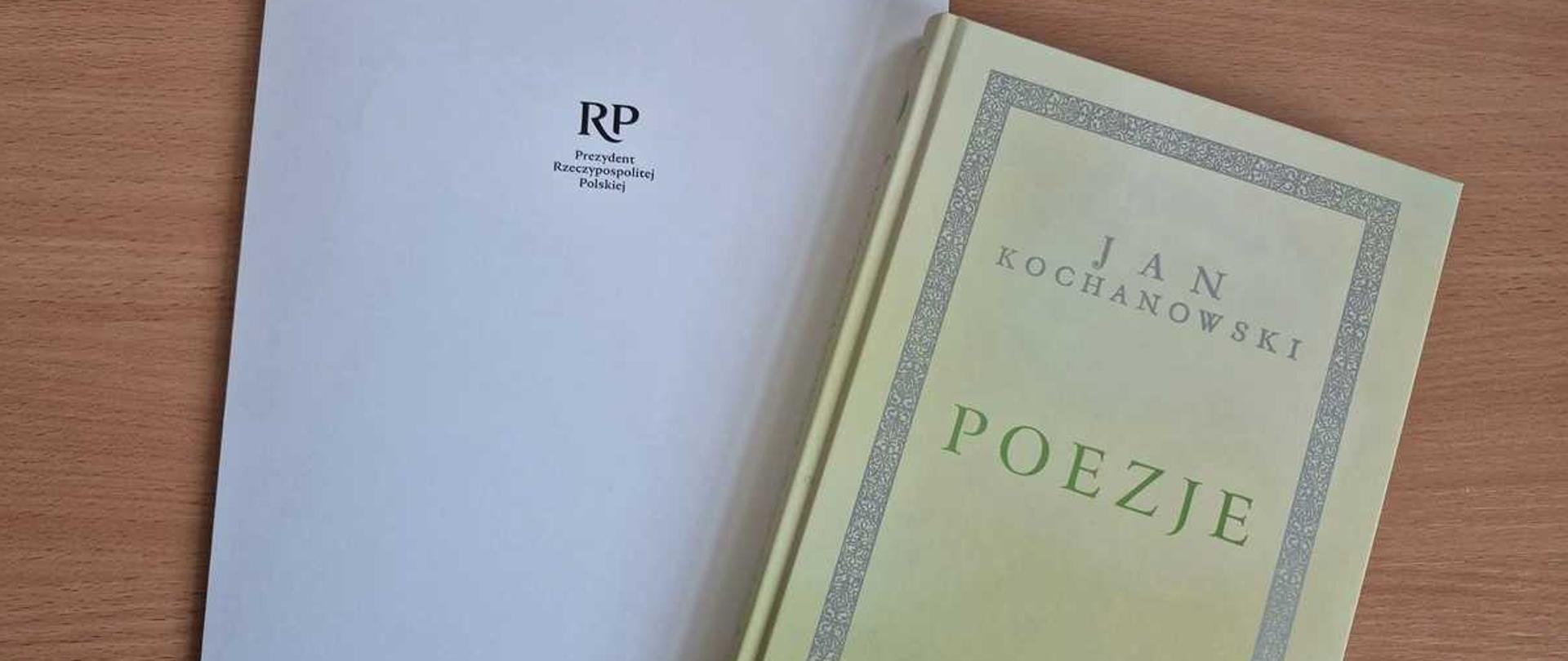 Obraz wyszukiwany wizualnie
opis zdjęcia dla niewidomych
Zdjęcie przedstawia dwa przedmioty leżące na drewnianej powierzchni.
Po lewej stronie znajduje się biały lub jasnoszary folder, na którym widnieje logo RP oraz tekst: ""Prezydent Rzeczypospolitej Polskiej"".
Po prawej stronie leży książka w miękkiej, jasnej (lekko żółto-zielonej) oprawie. Na okładce znajduje się ozdobna ramka, a wewnątrz niej tytuł: ""JAN KOCHANOWSKI"" oraz dużymi, zielonymi literami ""POEZJE"". W prawym dolnym rogu okładki znajduje się małe czerwone logo z napisem ""NARODOWE CZYTANIE"".