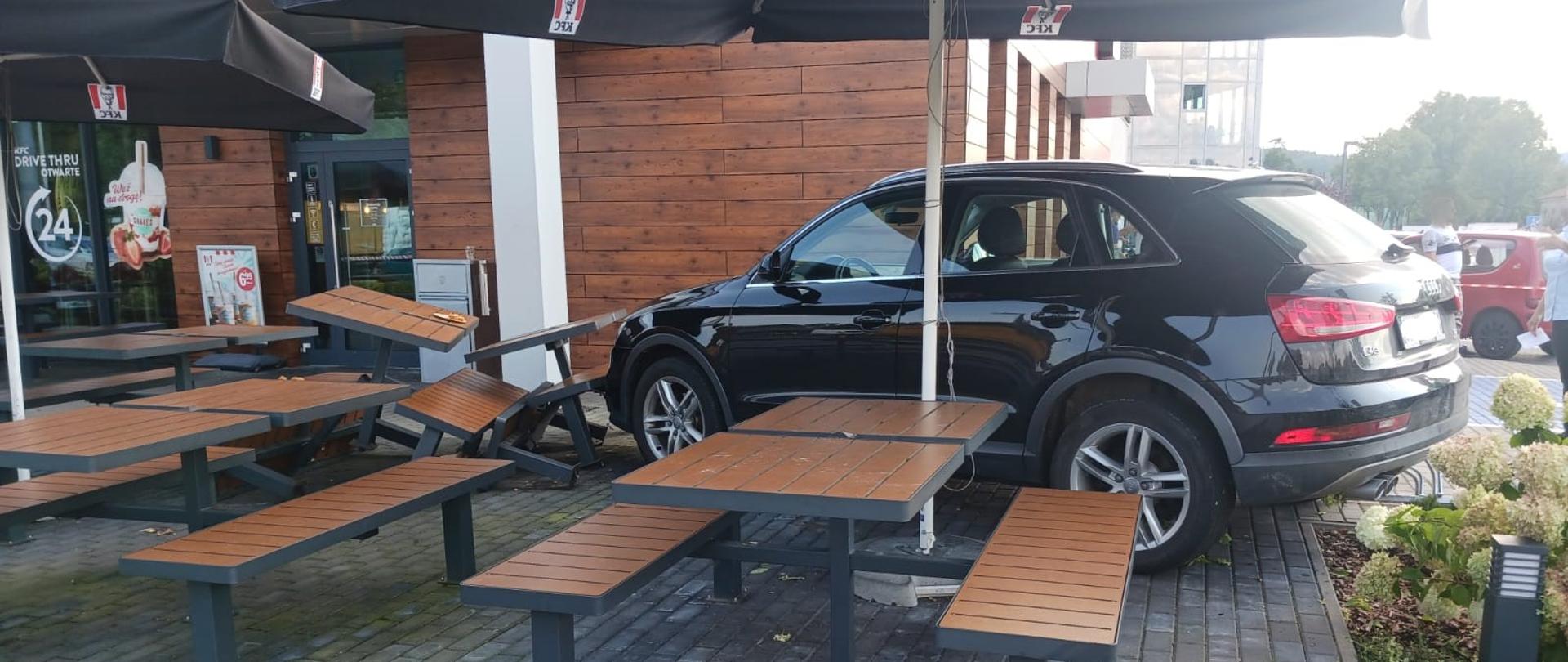 Widok z boku. Samochód osobowy w ogródku restauracyjnym. Przed samochodem przewrócone stoliki i ławki. W tle budynek restauracji. 