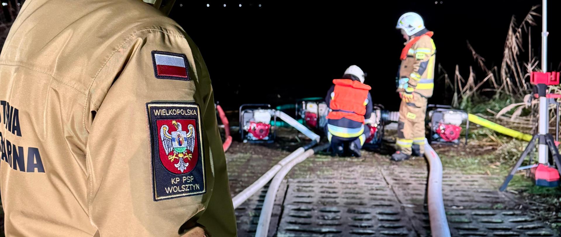 Nocna scena działań strażackich w terenie leśnym lub na nieutwardzonym placu. Widać rozstawione samochody pożarnicze z włączonym oświetleniem masztów, które intensywnie oświetlają miejsce pracy. Na pierwszym planie leżą rozwinięte linie wężowe, a strażacy przygotowują lub obsługują sprzęt. W tle dostrzegalne są krople deszczu lub śniegu odbijające światło reflektorów.