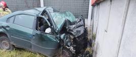 Zdjęcie przedstawia samochód osobowy po wypadku samochodowym, który wjechał w betonowy słup wiaduktu drogowego. Przód pojazdu jest tak mocno zniszczony, że znajduje się na wysokości przedniej szyby. W tle widać strażaka.