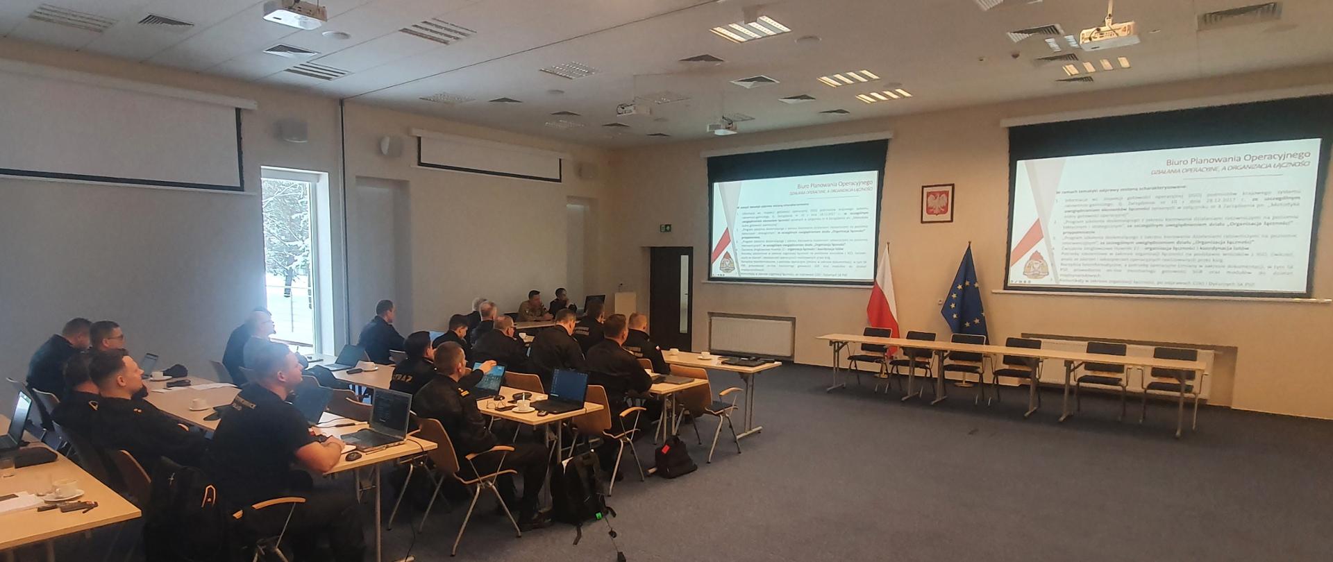 W ławkach siedzą uczestnicy warsztatów, przed nimi dwa ekrany z wyświetlonymi slajdami z zakresu omawianych zagadnień