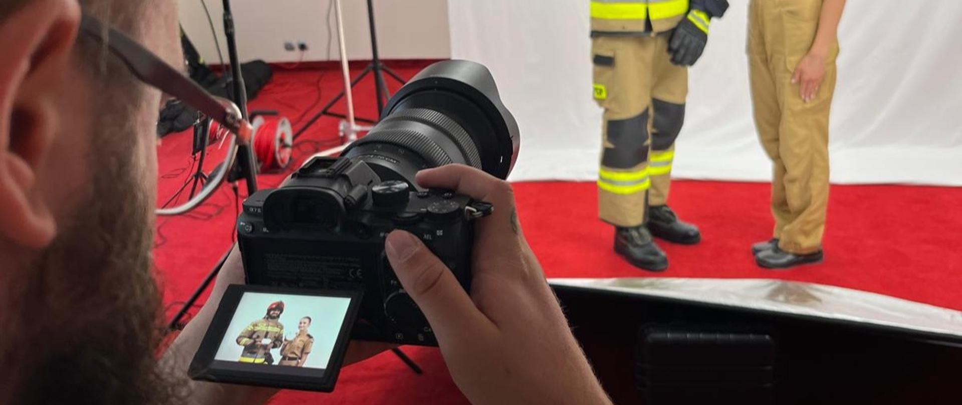 Z lewej strony fotograf z aparatem, przed nim para strażaków pozujących z czujkami. Czerwony dwan.