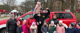 Zdjęcie grupowe dzieci i strażaka na tle samochodu pożarniczego typu pickup. Strażak trzyma na rękach jedno z dzieci.