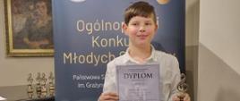 Uczeń stoi na tle baneru XXI Ogólnopolskiego Konkursu Młodych Solistów w Jaworznie, trzyma dyplom I miejsca i statuetkę w kształcie klucza wiolinowego