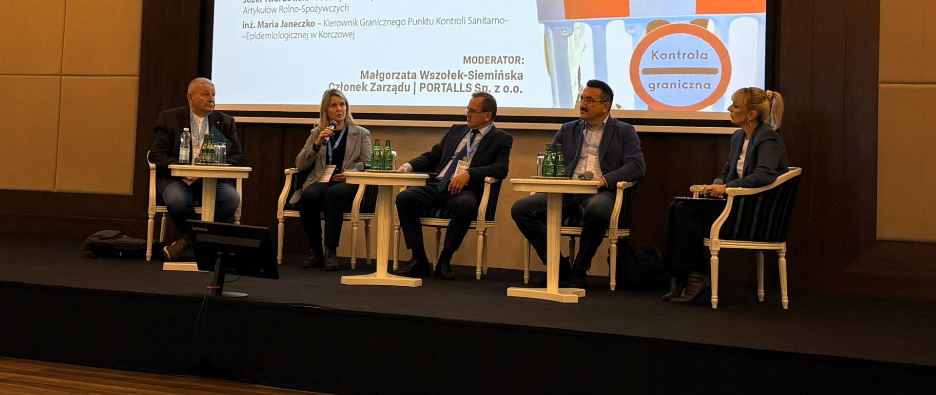 Grupa ludzi siedzi na krzesłach które znajdują się na scenie. Za nimi wyświetlana jest prezentacja. Ubrani są na elegancko, w garnitury.