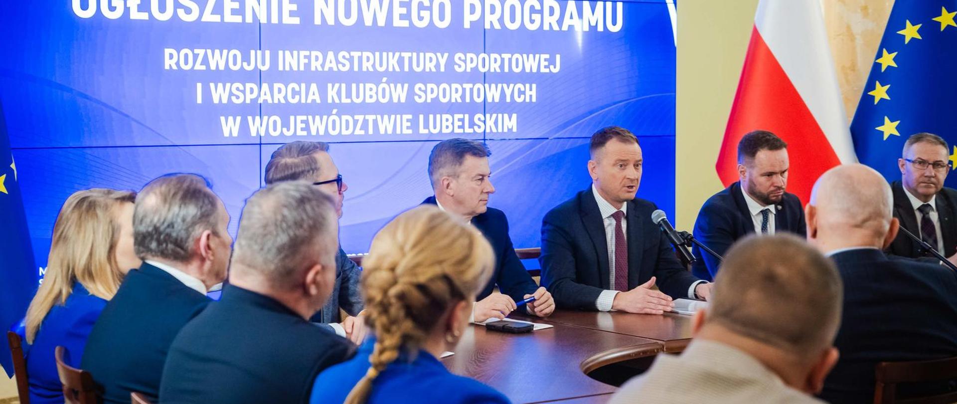 Minister Sławomir Nitras siedzi za stołem i mówi do mikrofonu. Po jego obu stronach siedzą inni ludzie. Za jego plecami prezentacja z napisem "Ogłoszenie nowego programu". Z boku flagi Polski i UE.