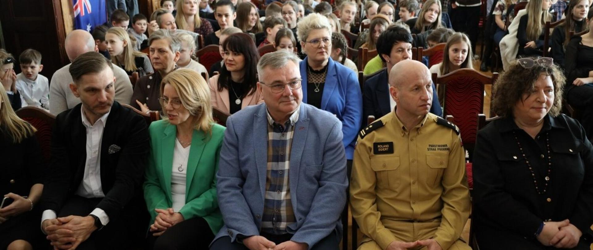 Na oświetlonej sali siedzi w rzędach grupa kobiet i mężczyzn, pośrodku siedzi mężczyzna w żółtym mundurze Państwowej Straży Pożarnej 