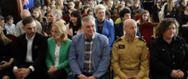 Na oświetlonej sali siedzi w rzędach grupa kobiet i mężczyzn, pośrodku siedzi mężczyzna w żółtym mundurze Państwowej Straży Pożarnej 